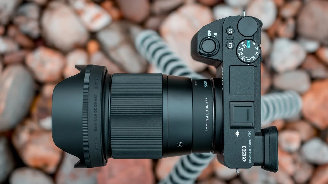 Об'єктив Sigma 16mm f/1.4 DC DN Contemporary встановлений на чорну бездзеркальну камеру, вид зверху, видно широке кільце фокусування та маркування різьби під світлофільтр діаметром 67 міліметрів на фоні розмитого каміння.