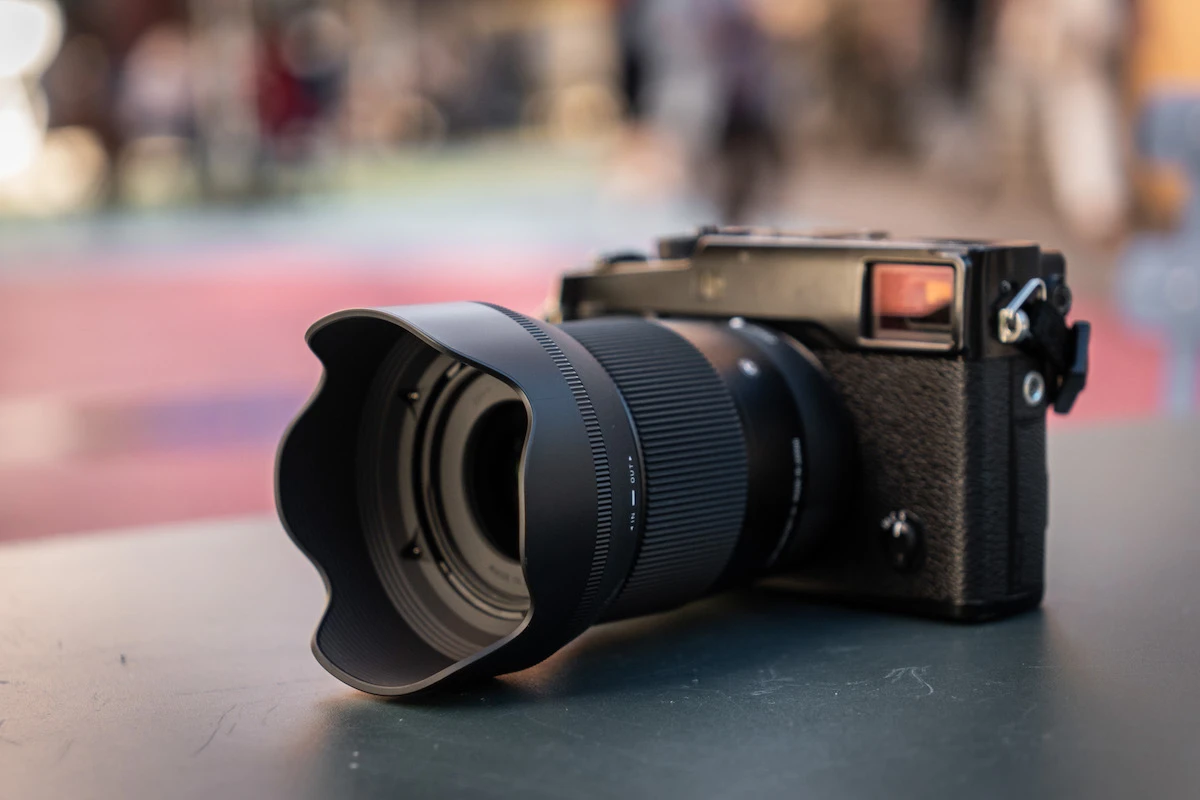 Компактна чорна фотокамера формату APS-C стоїть на столі з прикріпленим об'єктивом Sigma, на об'єктив надіта пелюсткова бленда, фоном служить сильно розмита вулиця міста з м'яким денним світлом.